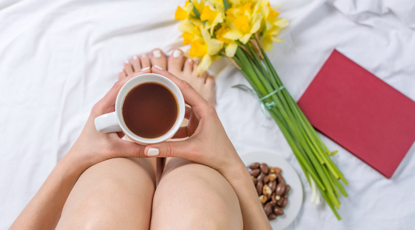 Feng-Shui-Mindfulness-for-the-home--Equate A woman sits on her bed with a cup of coffee, snacks a red book and daffodils.