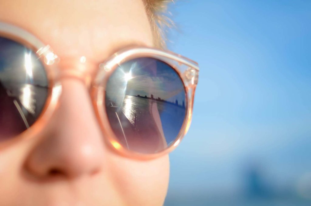 Woman gazing into distance, sunglasses on with reflection of sun and city across the water
