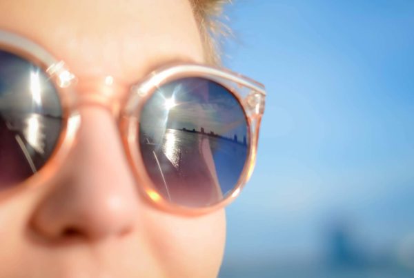 Woman gazing into distance, sunglasses on with reflection of sun and city across the water