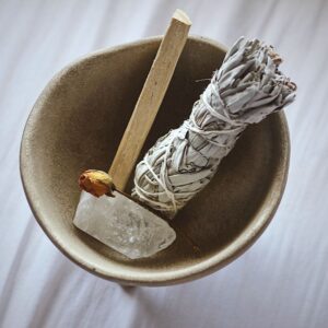 White sage bundle, palo santo rose and quartz all arranged in a bowl on a white sheet background. Image by Karly Jones on UnSplash