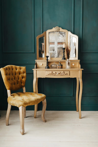 A golden vintage chair and wooden dressing table and mirror stand against a darkgreen painted wall